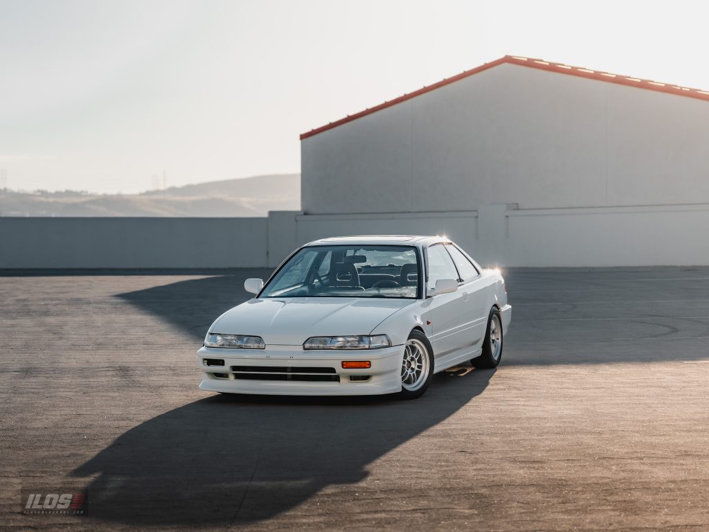 White early-90s Acura Integra coupe with aftermarket wheels parked in a lot, photographed from the front three-quarter angle